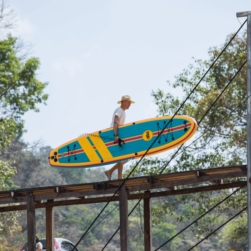 A man carrying a POP Yacht Hopper 11' Inflatable Touring Paddle Board SUP – Turq/Neon/Pink on a bridge