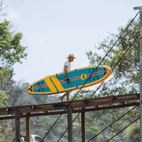 Thumbnail for A man carrying a POP Yacht Hopper 11' Inflatable Touring Paddle Board SUP – Turq/Neon/Pink on a bridge