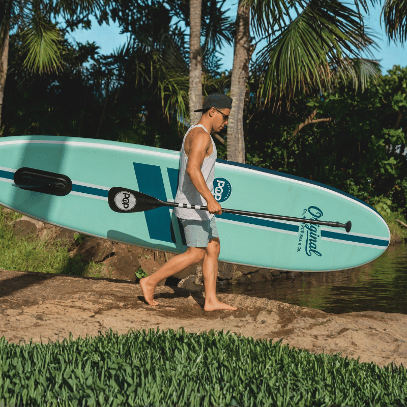 A man carrying a POP Yacht Hopper 11' Inflatable Touring Paddle Board SUP with Cooler Mount – Mint/Teak/Blue